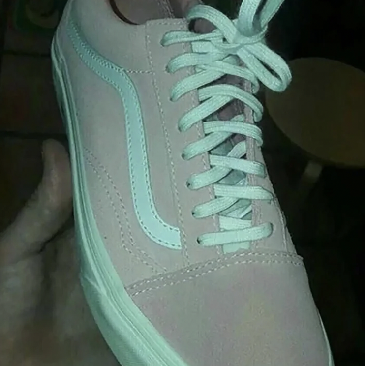 A hand holding a sneaker under dim light, causing debate over its color. The shoe appears to be a light pink or gray with light blue or white accents and white laces, creating an optical illusion of different possible color combinations.