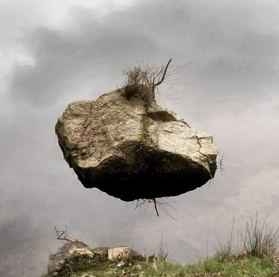 A large rock appears to be floating in mid-air against a cloudy sky background. Sparse grass and small bushes are growing on top of the rock, adding to the surreal scene. At the bottom, more grass can be seen, suggesting the rock is reflecting off water.