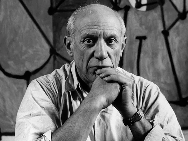 A black and white photo of an older man with a bald head and expressive eyes, resting his chin on his clasped hands. He wears a light-colored shirt and a wristwatch, with an abstract painting in the background.