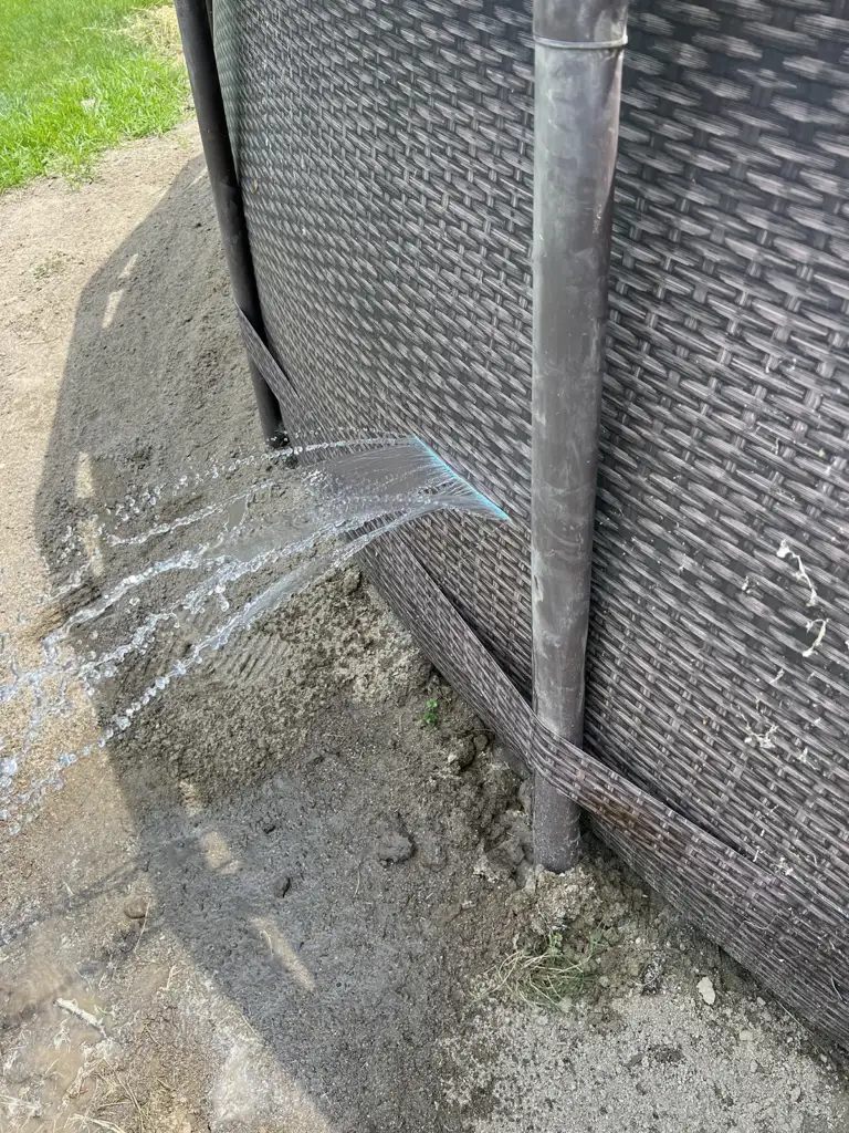 Water is spraying out from a hole in the side of an above-ground swimming pool, splashing onto the dirt and ground below. The pool exterior has a wicker-like design and is supported by metal poles.