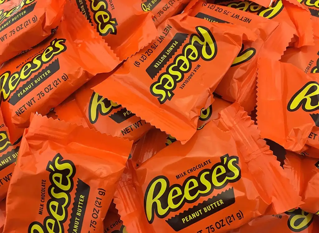 A pile of individually wrapped Reese's Peanut Butter Cups is shown. The wrappers are bright orange with the Reese's logo prominently displayed in yellow and black, indicating the candy contains milk chocolate and peanut butter.