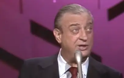 A man with gray hair slicked back is speaking into a microphone. He is wearing a dark suit jacket, a white shirt, and a maroon tie. The background is pink with black geometric shapes. The man appears to be on stage.