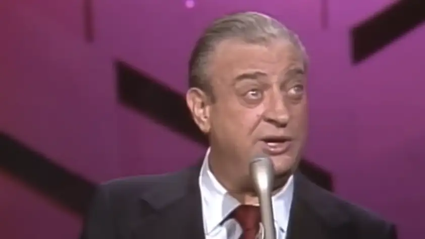 A man with gray hair slicked back is speaking into a microphone. He is wearing a dark suit jacket, a white shirt, and a maroon tie. The background is pink with black geometric shapes. The man appears to be on stage.