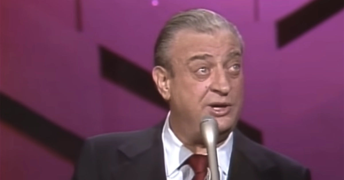 A man with gray hair slicked back is speaking into a microphone. He is wearing a dark suit jacket, a white shirt, and a maroon tie. The background is pink with black geometric shapes. The man appears to be on stage.