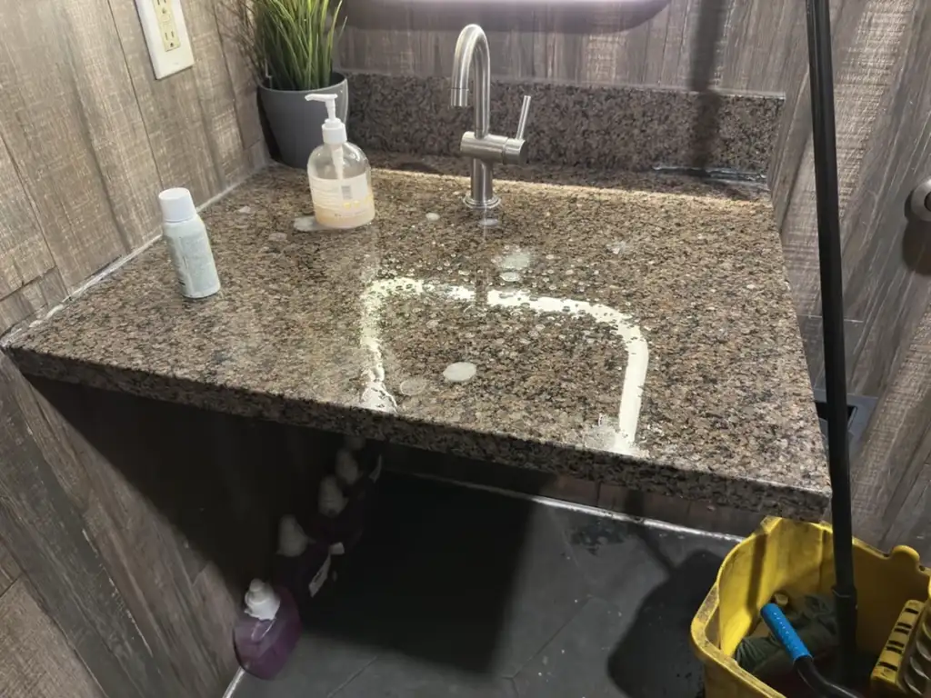 A bathroom sink with a granite countertop is shown. The sink has some soap residue and is surrounded by a soap dispenser, a small bottle, and a potted plant. Cleaning supplies, including a yellow mop bucket, are visible beneath the sink.