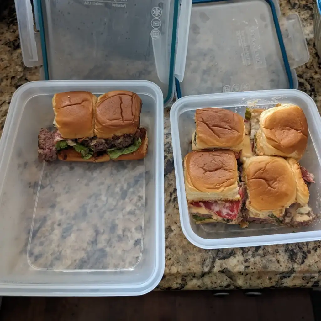 Two plastic containers on a granite countertop, each holding three sizable slider sandwiches made with dinner rolls. The sliders contain visible layers of meat, cheese, and other toppings. One container is open, while the other has its lid placed beside it.