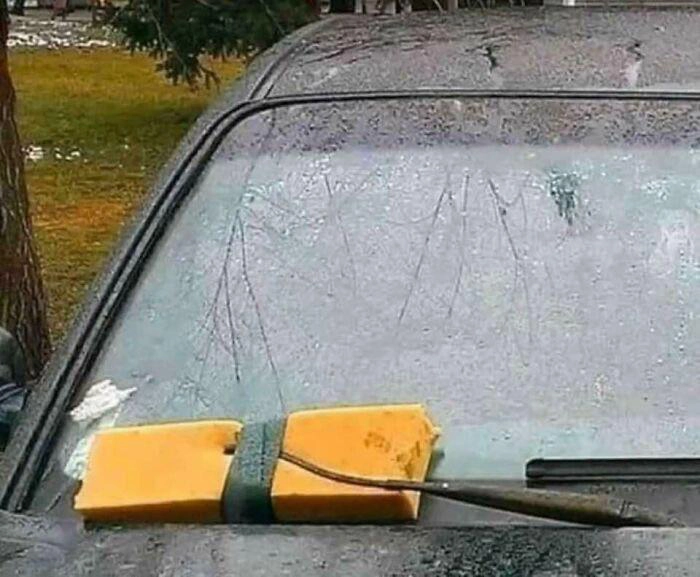 A car windshield with a large yellow sponge attached to the wiper arm with a strap instead of a traditional wiper blade. The car and surrounding area appear wet as if it has recently rained. Trees and grass are visible in the background.