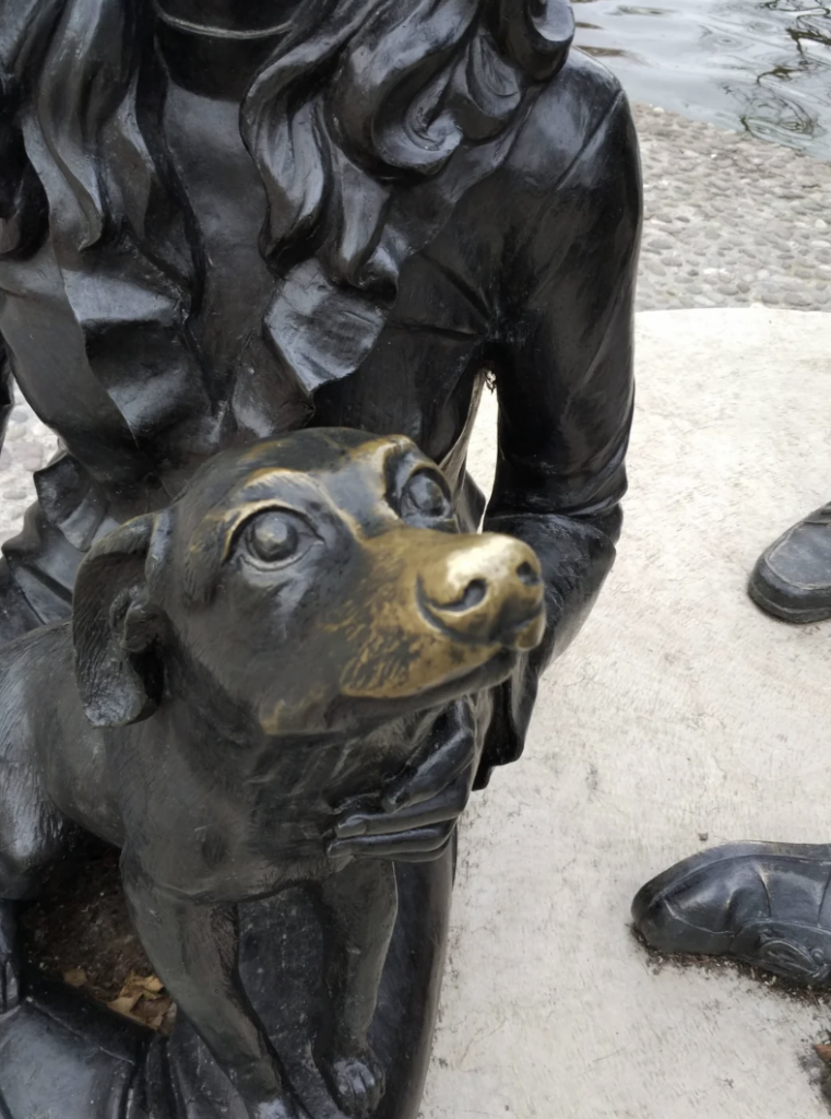 A bronze statue of a person with curly hair kneeling next to a dog with a golden snout. The dog's face is tilted upwards, and the hands of the person are gently holding it. The statue is set on a stone surface with pebbles and a water feature in the background.