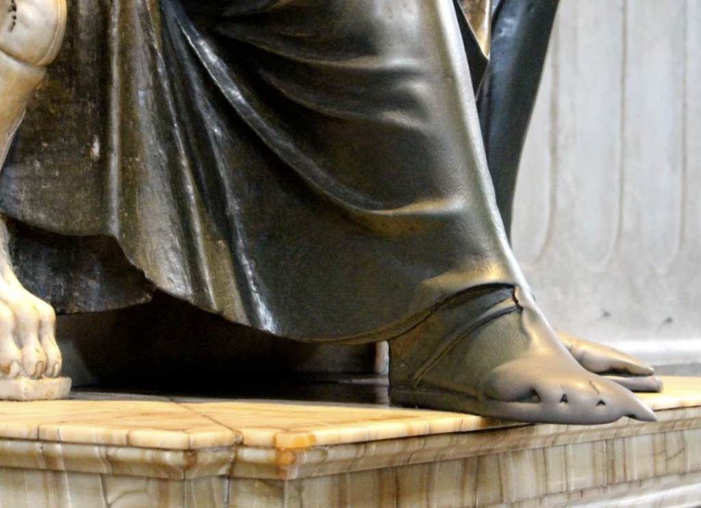 Close-up of a statue showing the lower part of a seated figure wearing a draped robe and sandals with lion-like claw details. The figure's foot rests on a polished stone base, and part of a lion's paw is visible next to the foot.