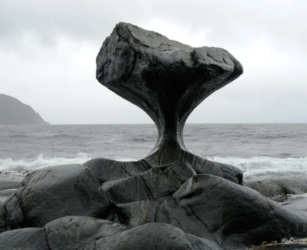 A unique rock formation known as Kannesteinen, located in Norway, stands on the shore with its base eroded by waves, creating a mushroom-like shape. It is surrounded by large, smooth rocks and an overcast sky above the ocean in the background.