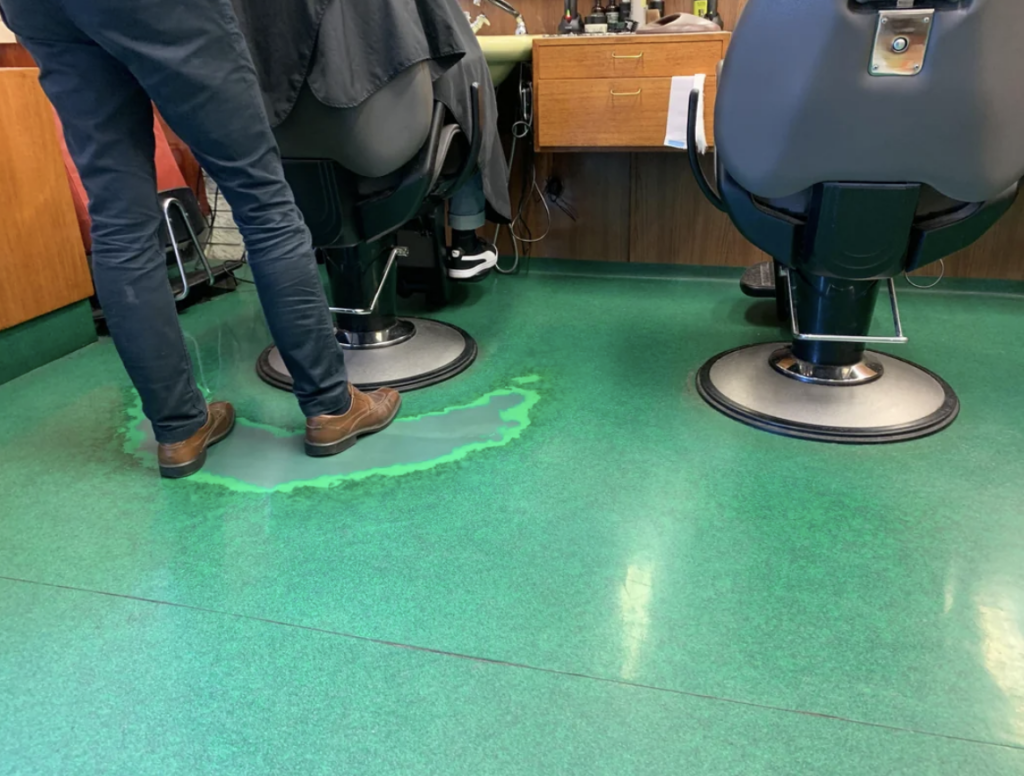 A person stands on a green floor that has a noticeable spill or wet stain near their feet. Two barber chairs are positioned nearby, each facing a small wooden counter against the wall. The scene appears to be inside a barbershop.