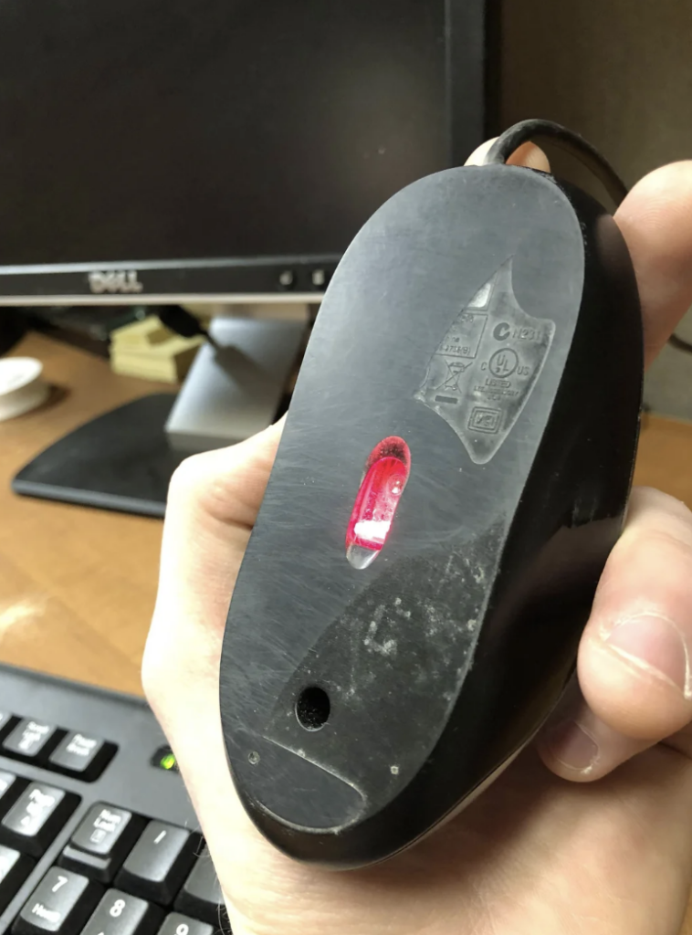 A hand holding a black computer mouse upside down, showing the bottom side with a red optical sensor illuminated. The mouse appears slightly worn, with visible scratches. There is a computer monitor and a keyboard in the background on a wooden desk.