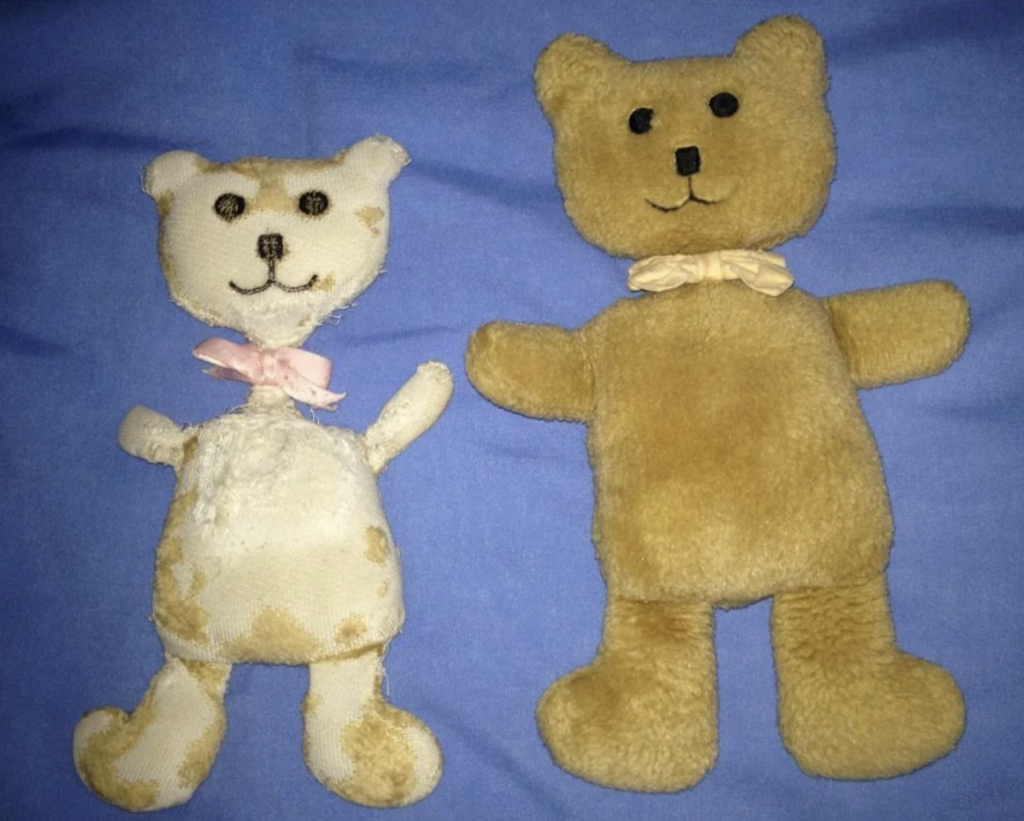 Two worn teddy bears lying on a blue surface. The bear on the left is white with brown spots and a pink bow, while the bear on the right is light brown with a cream-colored bow. Both bears have black button eyes and stitched smiles.