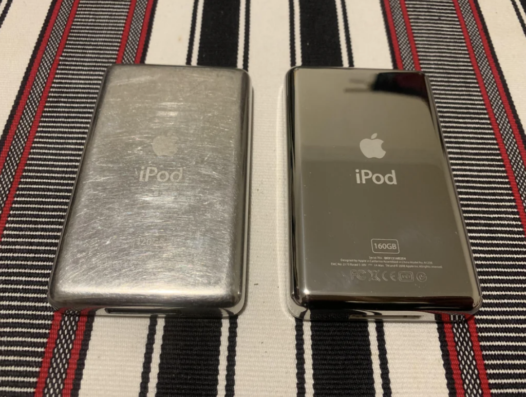 Two iPods placed side by side on a striped fabric surface. The one on the left has a brushed metal back with visible wear, while the one on the right has a shiny, polished back with the "iPod" logo and "160GB" capacity mark clearly visible.