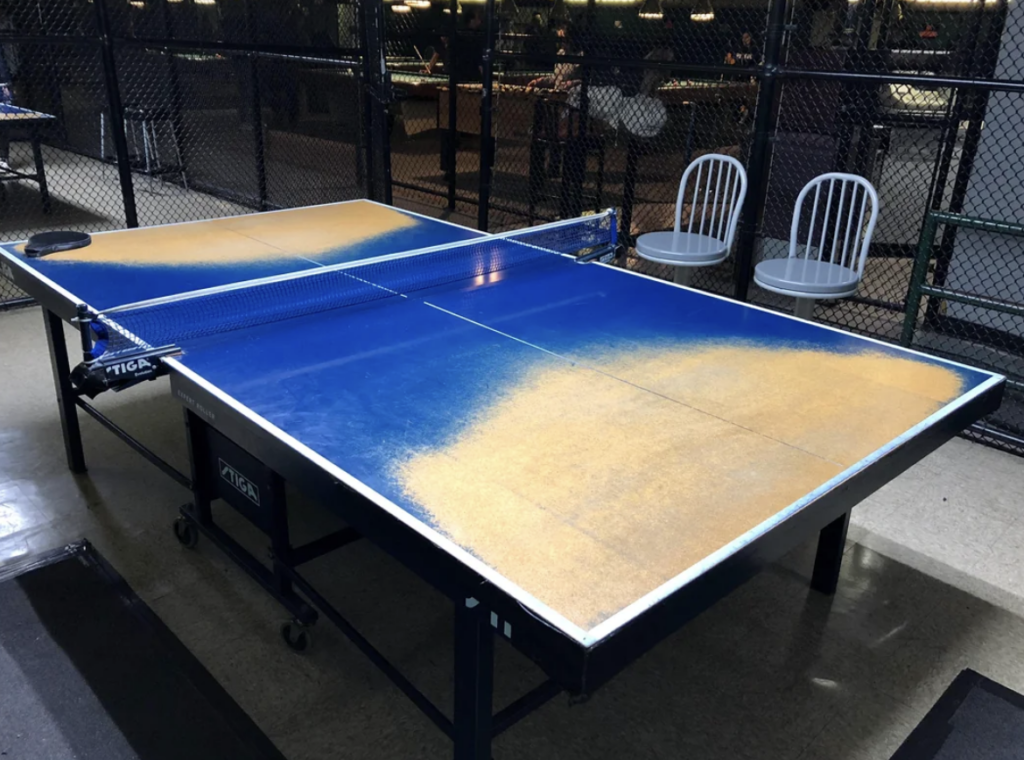 A ping pong table with a blue and yellow gradient design, located indoors near a chain-link fence. Two white chairs are placed side by side next to the table. The area appears to be in a recreational or game room.