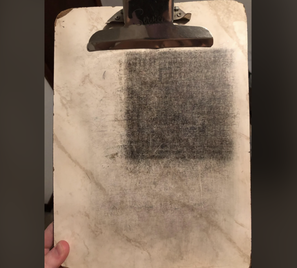 A worn clipboard with a large metal clip holds a piece of paper that has a dark, rectangular area smudged with pencil marks, blending into a lighter background. A person's hand is visible, holding the clipboard at the bottom.