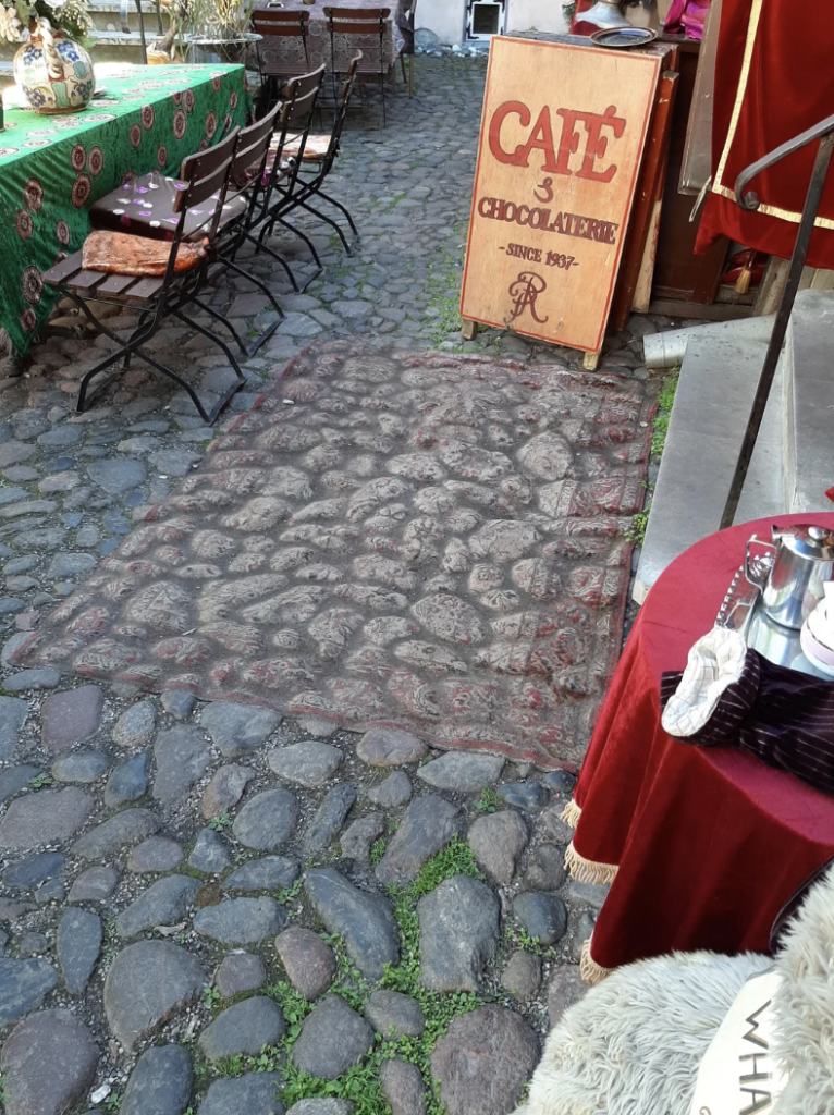 A cobblestone alley features an outdoor cafe with tables and chairs adorned with patterned green tablecloths. A wooden sign positioned near the entrance reads "Café Chocolaterie, Since 1892" in red letters. Nearby is another table decorated with a red cloth.