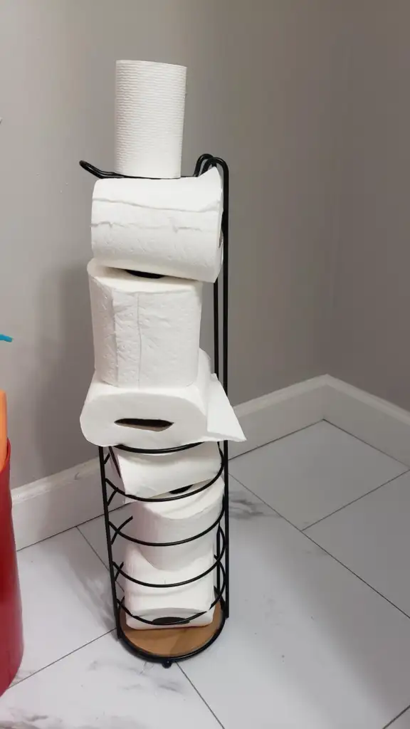 A black metal holder is stacked with seven rolls of toilet paper. It stands on a white tiled floor in a corner against gray walls. The bottom roll appears to be slightly unrolled, and a red container is partially visible to the left.