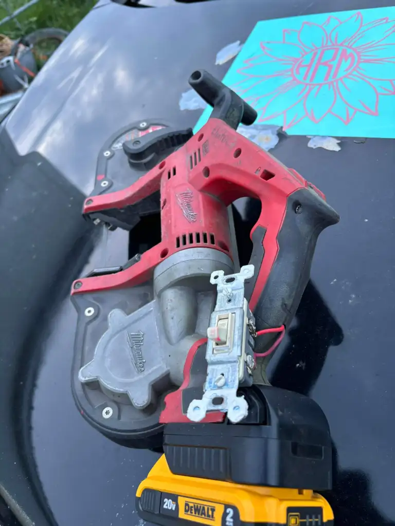 A red Milwaukee power tool and a DeWalt 20V battery lie on a dark surface. A single light switch is placed on the battery. In the background, there is a decorative blue sheet with a flower-like design.