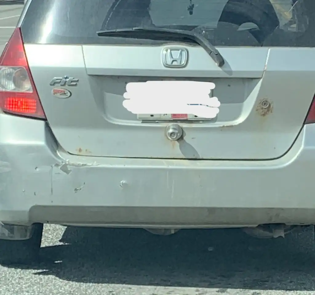 A silver Honda Fit with a slightly dented and rusty rear bumper is stopped in traffic. The car also has visible rust near the license plate, which is obscured for privacy. The shadows suggest it is a sunny day.