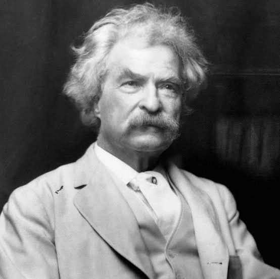An elderly man with white, curly hair and a thick mustache, dressed in a light-colored suit jacket over a high-collared shirt. He is looking slightly to the side with a serious expression. The background is dark and plain.