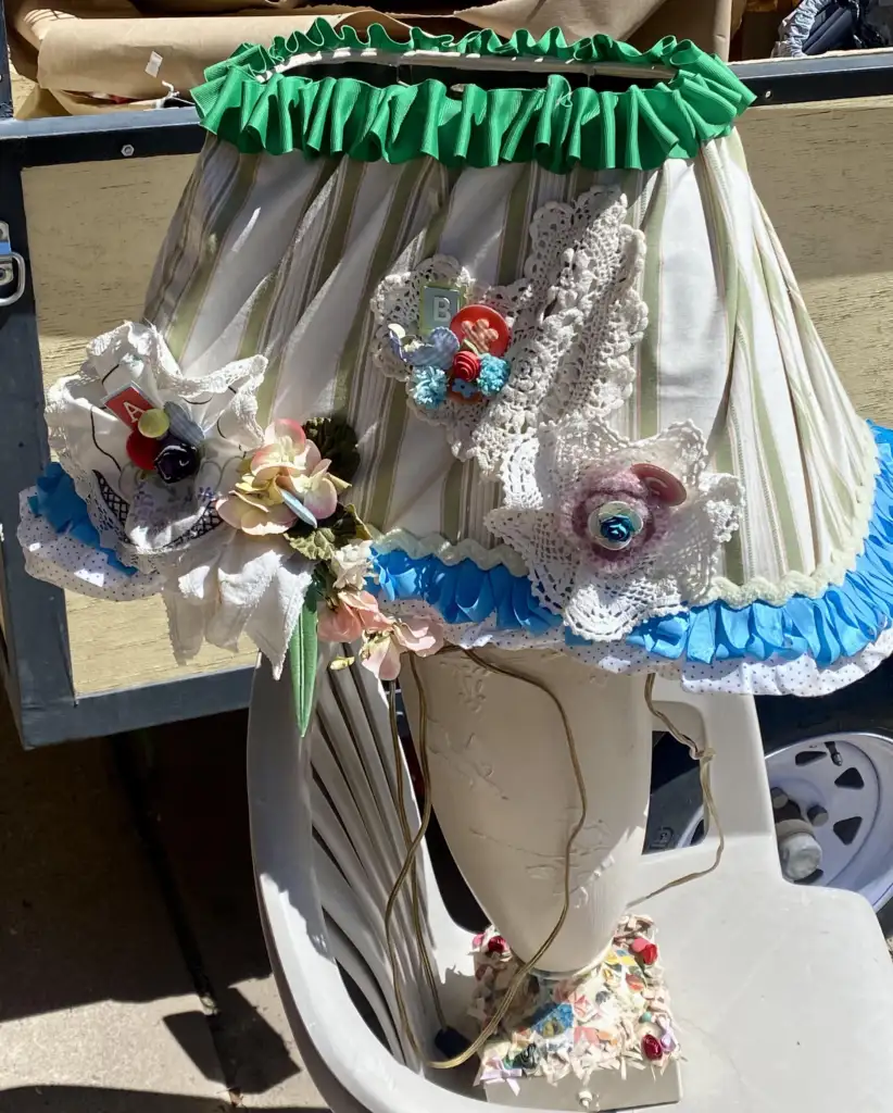 A whimsically decorated lampshade sits on a white chair. It features ruffled green and blue trim, striped fabric, and various attached ornaments including flowers, lace, and a small gnome figurine. The background shows part of a wooden structure and other miscellaneous items.