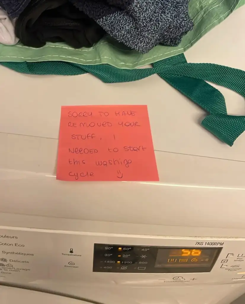 A handwritten note on a pink sticky note is placed on top of a white washing machine. The note reads, "Sorry to have removed your stuff, I needed to start this washing cycle :)". In the corner, a green and white bag and clothes can be seen.