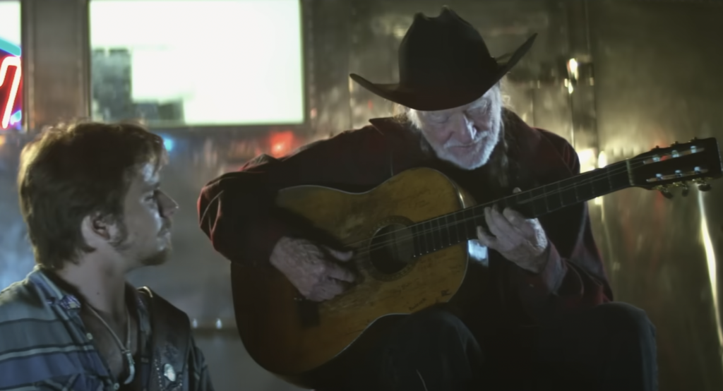 A still image of Willie Nelson playing guitar to his son. 