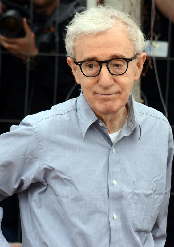 An older man with white hair and glasses, wearing a light blue button-down shirt, stands outdoors with people and a camera visible in the background.