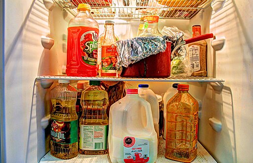 An open refrigerator displaying various items, including a red jug of V8 Splash juice on the top shelf, a foil-covered pot, condiments, and sauces. The bottom shelf holds a gallon of milk, cooking oil, juice bottles, and a container of iced tea.