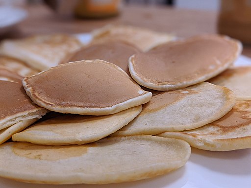 A close-up view of a stack of golden brown pancakes on a white plate. Some pancakes are slightly overlapping, showcasing their fluffy and smooth textures. The background is slightly blurred, putting focus on the appetizing pancakes.