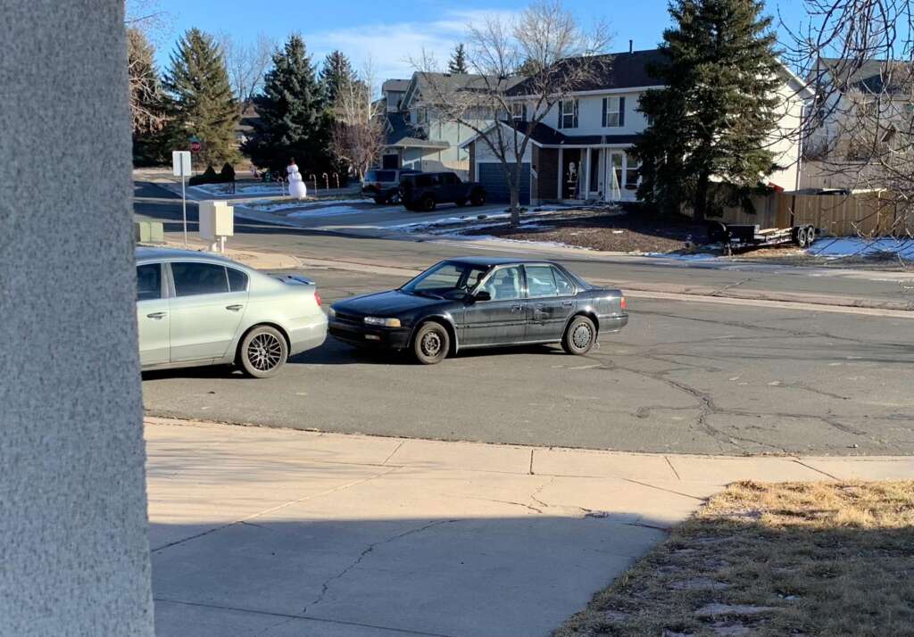 A black sedan is parked on the street next to a silver car on a sunny day in a suburban neighborhood. Snow is visible on parts of the ground, and there are houses, trees, and a snowman in the background.