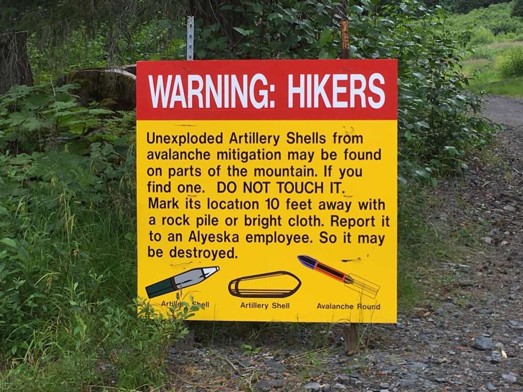 A large red and yellow sign warns hikers about unexploded artillery shells from avalanche mitigation on the mountain. The sign advises not to touch the shells, to mark their location, and to report it to an Alyeska employee. It features illustrations of shells and rounds.