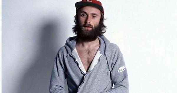 A bearded man wearing a gray zip-up hoodie and a red cap stands against a plain white background. His hoodie is partially unzipped, revealing part of his chest. He has a pensive expression and his hands are clasped in front of him.