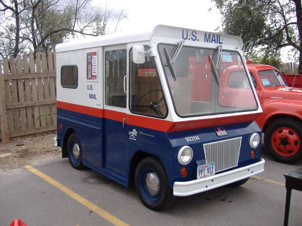 A vintage blue and white U.S. Mail truck is parked in an outdoor setting. The truck has a small, boxy design with "U.S. MAIL" written on the side and a postal emblem on the door. It is parked on a road with a wooden fence and trees in the background.