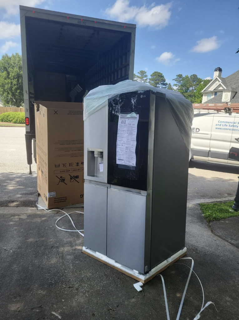 A large stainless steel refrigerator is wrapped in protective materials sitting outside on a driveway with a moving truck in the background. An empty cardboard box stands nearby. The scene appears to be a delivery or move-in process in a residential neighborhood.
