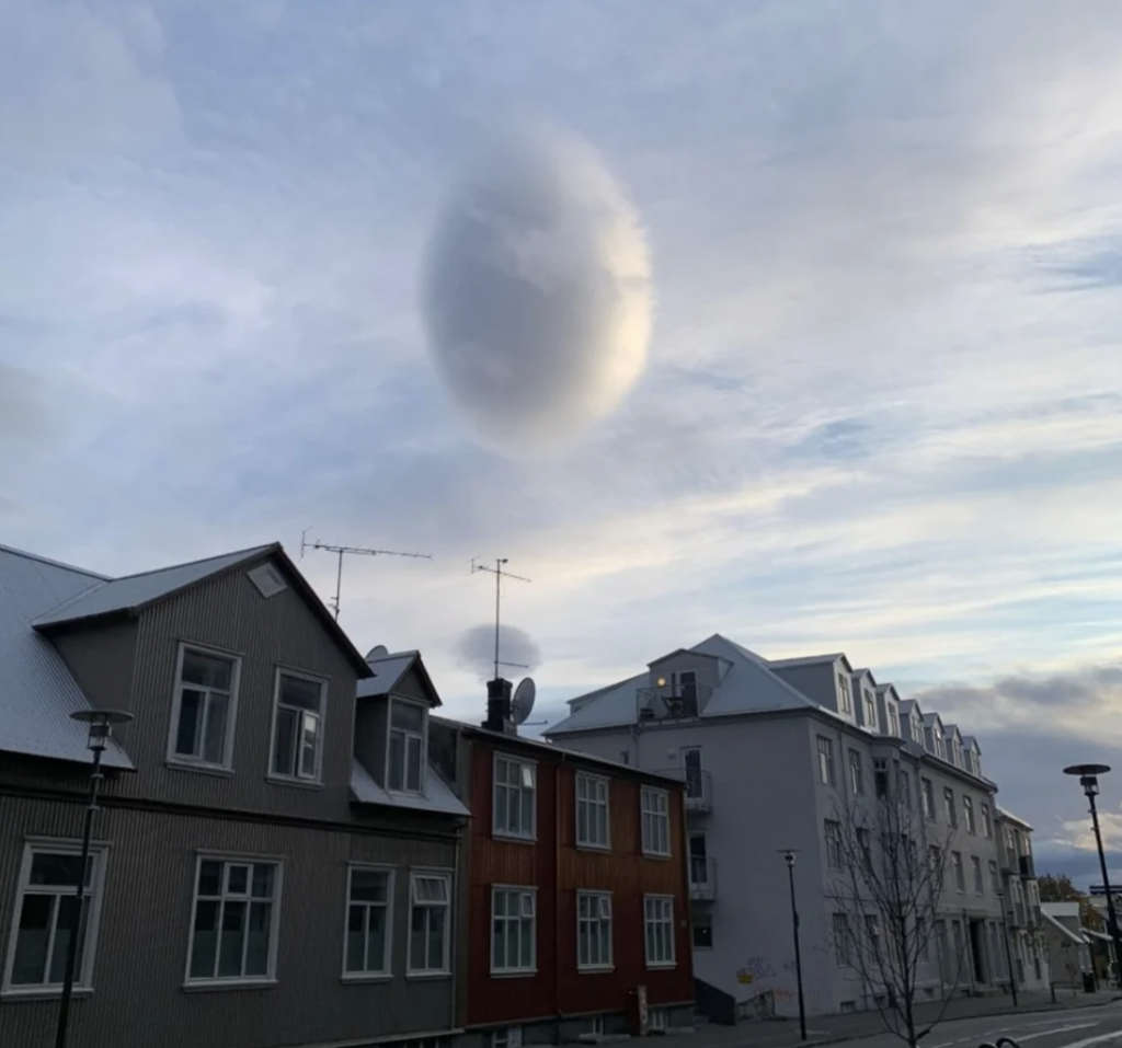 A scene depicting a cloudy sky above a residential area with several buildings featuring wooden facades and chimneys. One particularly intriguing, circular cloud formation is visible in the sky, appearing like a large, isolated puff of white in the otherwise streaky clouds.