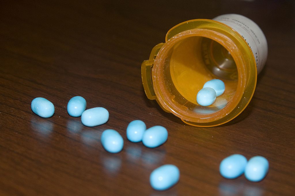 A prescription bottle with a yellow cap is tipped over on a wooden surface, spilling small, oval-shaped, blue pills. Some pills remain inside the bottle while others scatter around it in various directions.