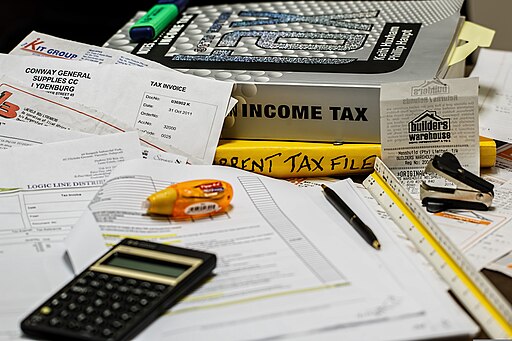 A cluttered desk with tax documents, a calculator, a large book labeled "Income Tax," receipts, and office supplies. The scene reflects the process of preparing for taxes, with various papers spread out and a focus on financial documents.