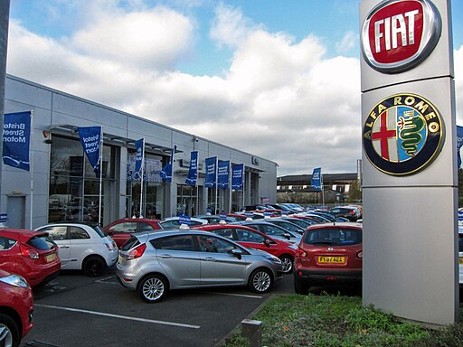 A Fiat and Alfa Romeo car dealership with a large sign displaying both brands' logos. The lot is filled with various cars, primarily in colors like red, white, and silver. Blue promotional banners are visible on the building in the background.