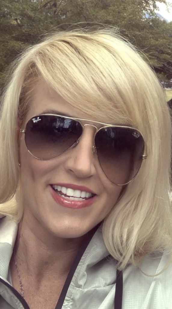 A woman with blonde hair and side-swept bangs is smiling at the camera. She is wearing large aviator sunglasses and a light-colored jacket. The background shows trees and greenery, suggesting an outdoor setting.