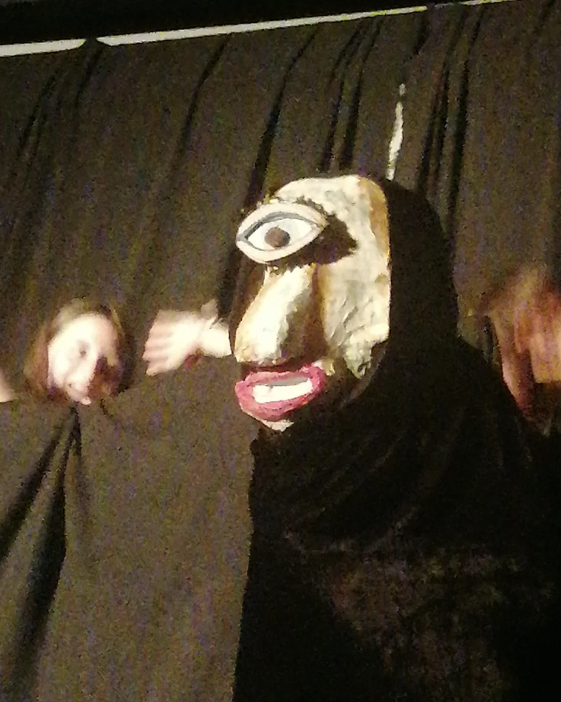 A person is performing on stage wearing a large mask with an exaggerated face and a single large eye in the center of the forehead. Two people with their arms wrapped in black fabric are in the background, creating a surreal and dramatic scene against a dark backdrop.