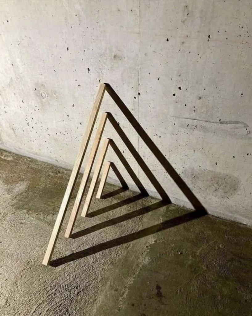 Four wooden triangular frames of varying sizes are nested concentrically, resting against a concrete wall in a dimly lit space. The careful alignment produces intriguing shadows, enhancing the geometric shape and adding depth to the minimalist arrangement.