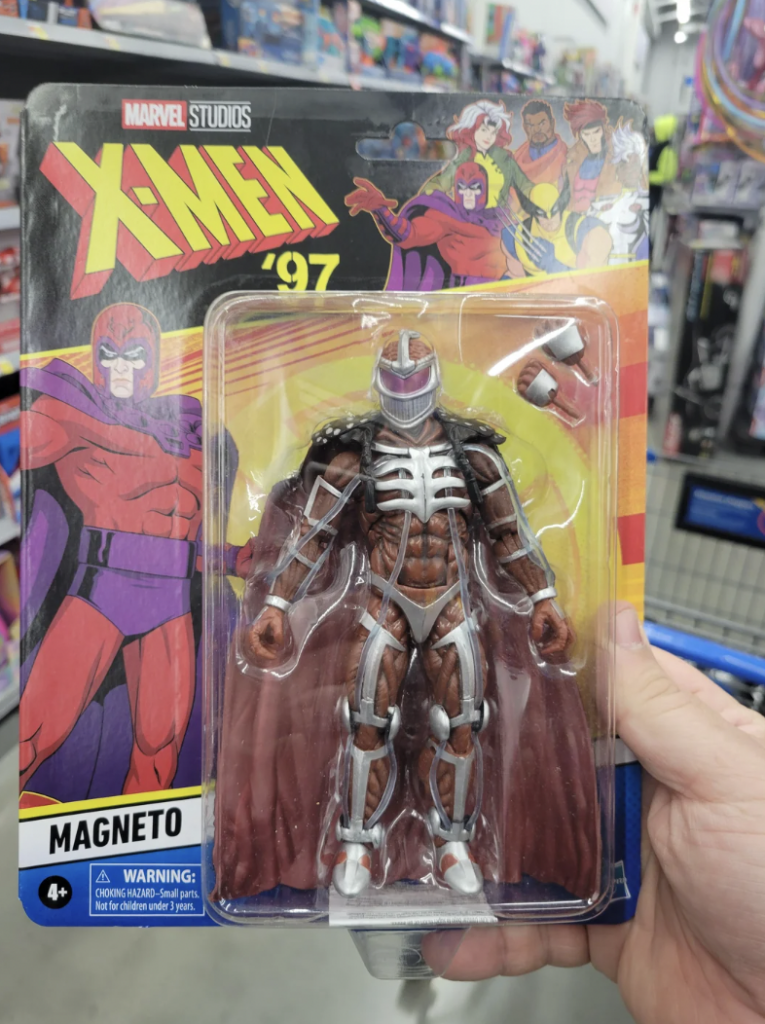 A hand is holding a packaged action figure of Magneto from the X-Men series. The packaging features colorful comic-style artwork of Magneto in a dynamic pose, with "X-Men '97" and "Magneto" prominently displayed. The figure has a metallic armor design and a red cape.