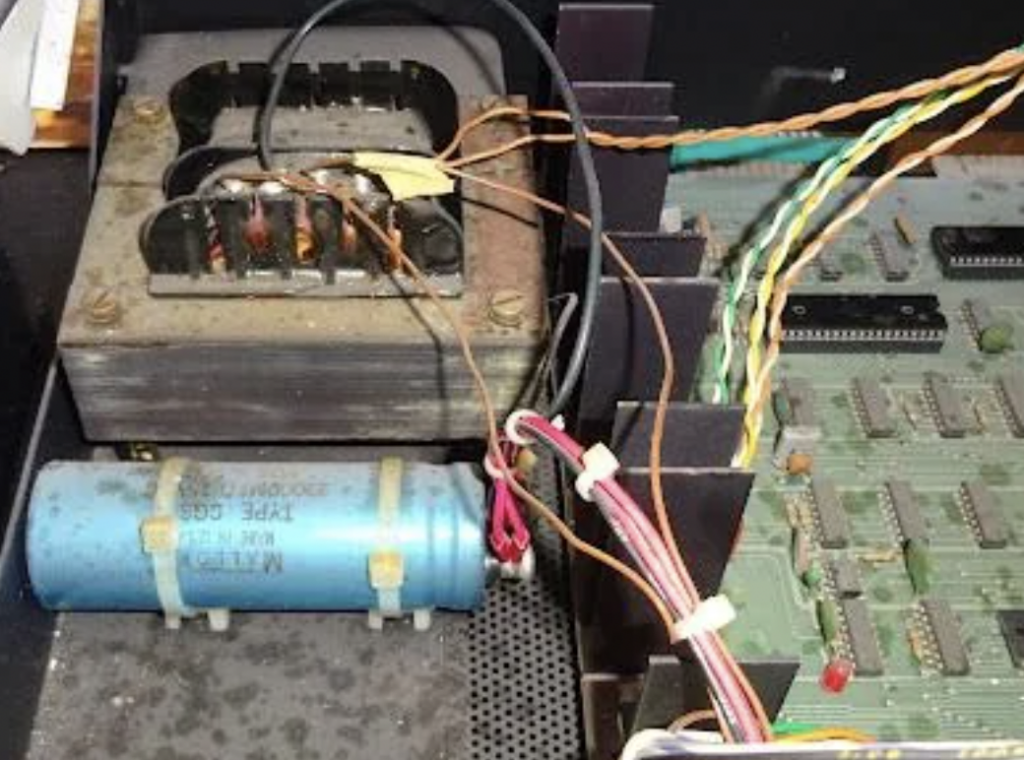 Close-up of the inside of an electronic device, showing a transformer, a blue capacitor, circuit board, and various connected wires. The components are placed within a metal and plastic housing, with visible connections, resistors, and integrated circuits.