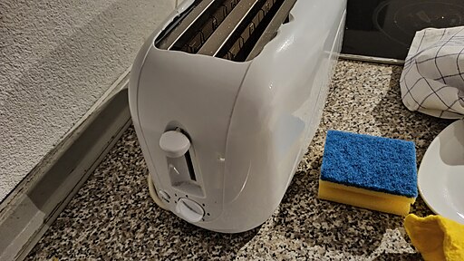 A white toaster sits on a speckled countertop beside a dual-sided sponge, one side yellow and the other blue. A white and blue dish towel and a white plate are partially visible on the right side of the frame.