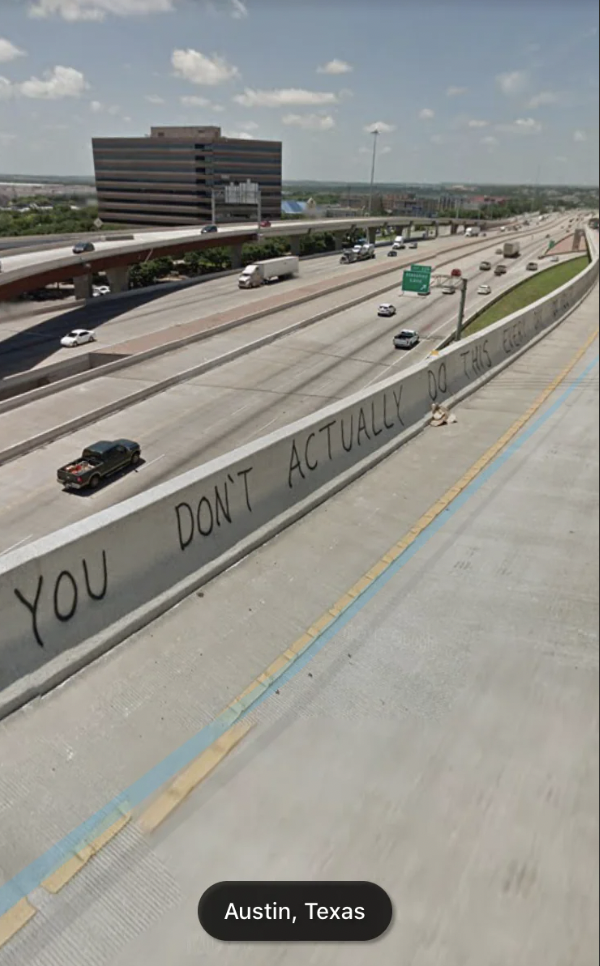 A highway in Austin, Texas, shows cars driving in both directions. The central concrete divider has graffiti that reads, "YOU DON'T ACTUALLY DO THIS FOR A LIVING, DO YOU?". The image is taken during the day under a clear sky.