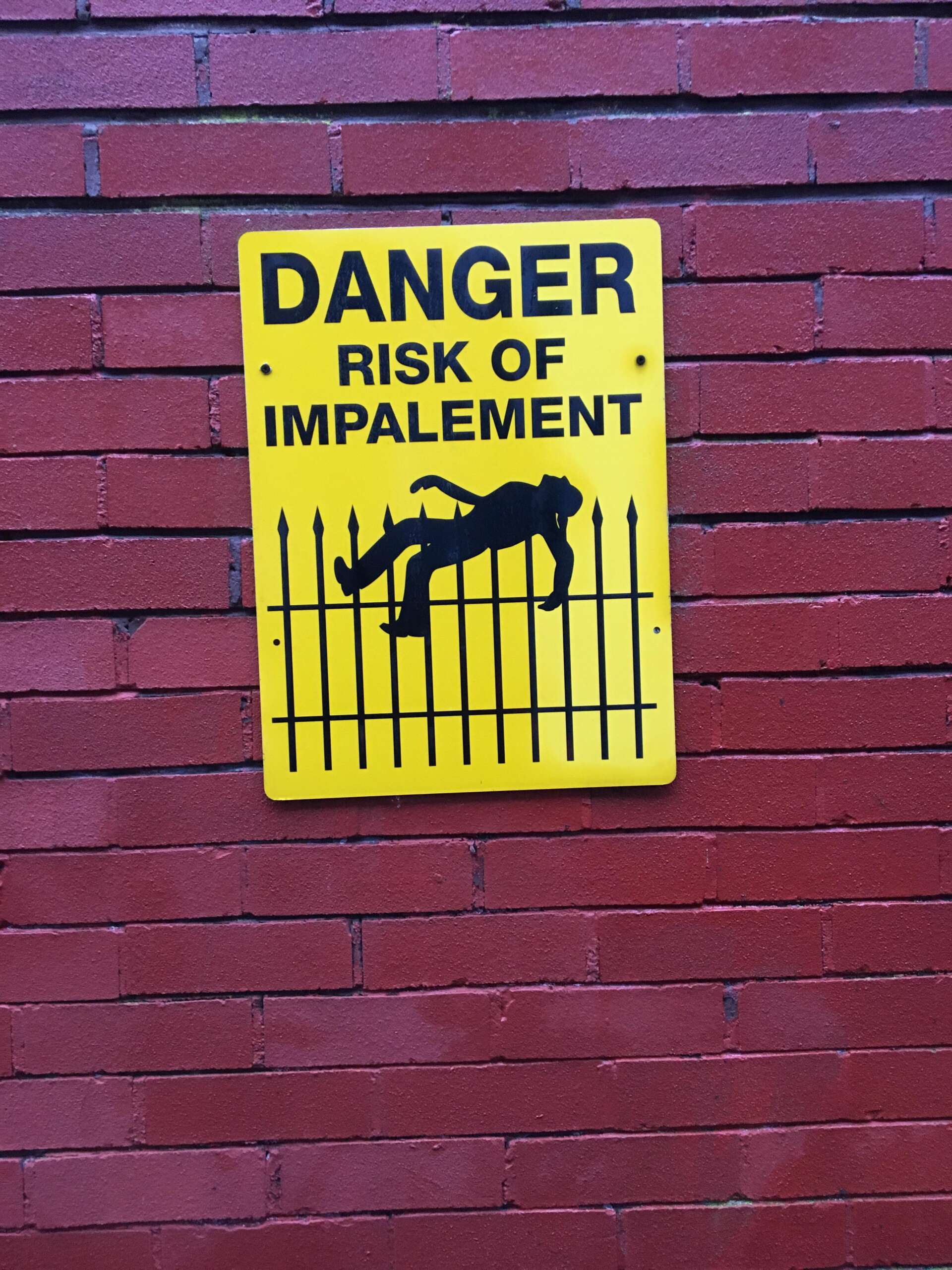 A yellow warning sign with black text that reads "DANGER RISK OF IMPALEMENT" is mounted on a red brick wall. The sign features an illustration of a person being impaled on a spiked fence.