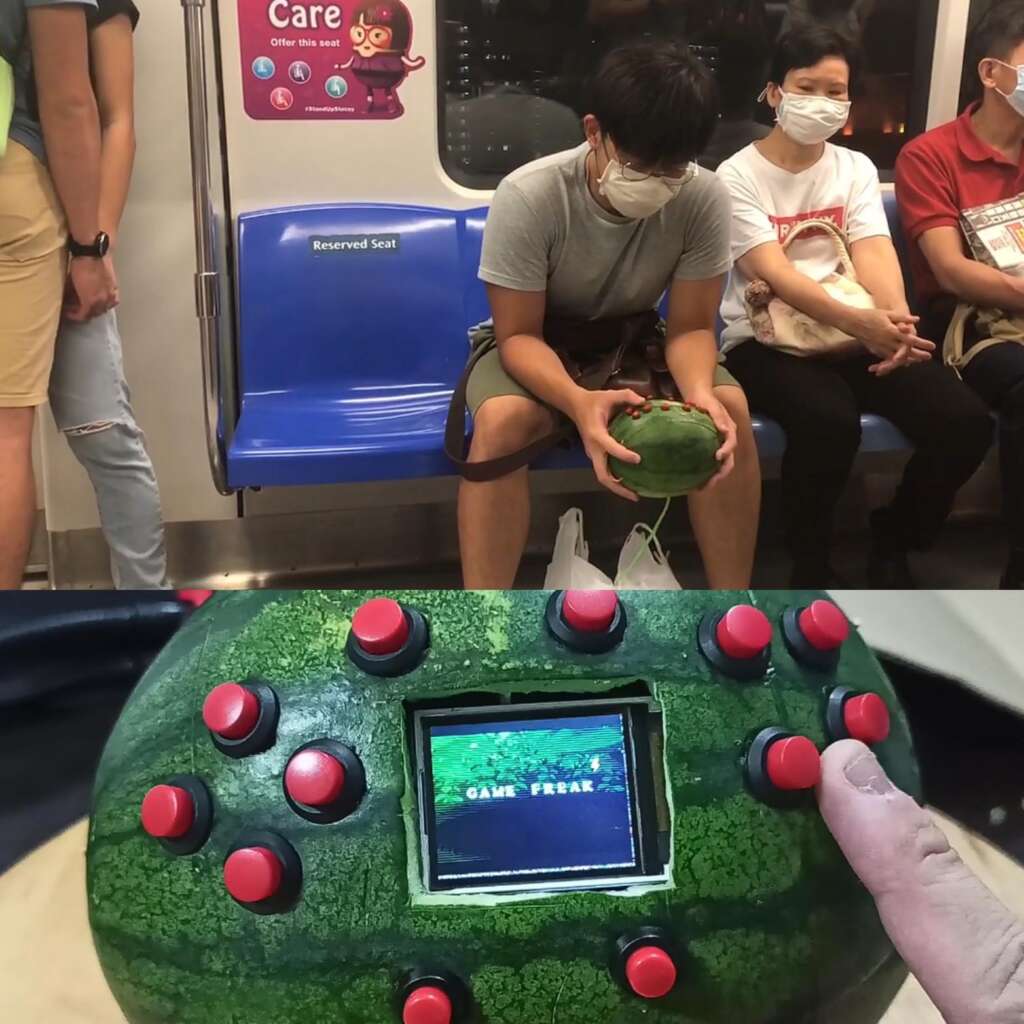 A person wearing a mask is sitting on a subway holding a watermelon modified into a portable gaming console. The watermelon has a small screen in the center and numerous red buttons around it. Two other masked passengers are seated nearby.