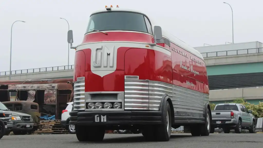 A streamlined, retro-futuristic red and white bus with a large "GM" emblem on the front, designed with an aerodynamically rounded nose. The bus is parked in an industrial area with old vehicles and a highway in the background.